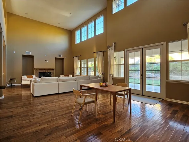 Dramatic two-story great room with soaring ceilings, rich hardwood floors, and expansive windows filling the space with natural light. Wall of windows and French doors open to the backyard patio, creating seamless indoor-outdoor flow. Spacious seating area centers around a cozy fireplace, ideal for entertaining or relaxing.