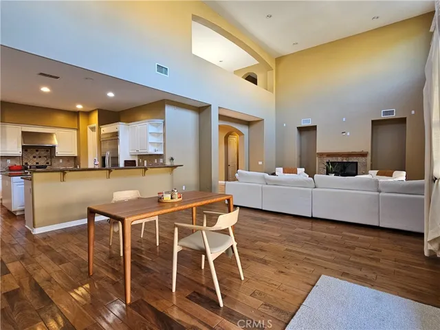 Open-concept great room with soaring ceilings, rich hardwood floors, and abundant natural light. Modern white kitchen offers granite counters, tile backsplash, and breakfast bar open to dining area. Spacious family room features large sectional seating and fireplace, ideal for entertaining and everyday living.