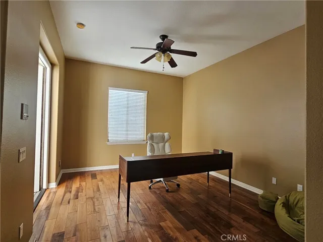 Spacious room featuring hardwood floors, neutral wall tones, a ceiling fan with light, and a large window providing natural light. Currently shown as a home office with direct access to the exterior.