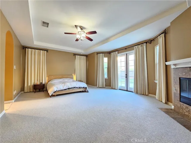 Discover the ultimate in comfort and convenience with this main-level primary bedroom suite. This expansive retreat features an elegant tray ceiling and a cozy stone fireplace, perfect for relaxing evenings. French doors open to the private outdoor space, filling the room with natural light. With a neutral palette, plush carpeting, and a ceiling fan, this functional and luxurious suite is designed for tranquility and easy living.