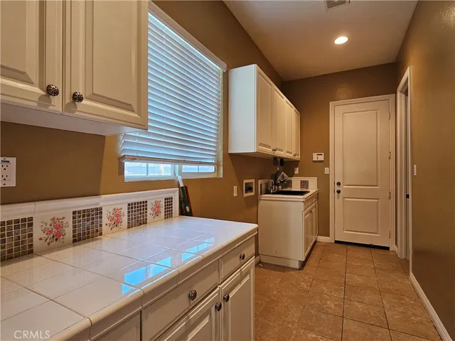 Dedicated laundry room with custom white cabinetry, utility sink, walk-in closet, and tiled counter space. Bright space with large window, faux wood blinds, and durable tile flooring.
