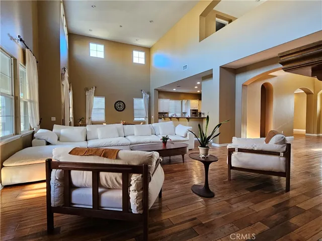 Spacious, light-filled great room with soaring ceilings, warm neutral walls, and rich hardwood floors. Oversized white sectional and lounge chairs create an inviting seating area, open to the kitchen and hallways through wide arches. Multiple tall windows and recessed lighting enhance the bright, airy feel of this open-concept living space.
