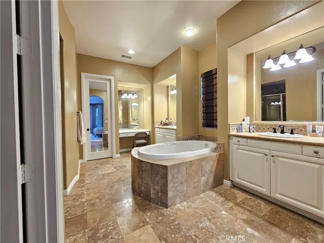 Indulge in this spa-inspired downstairs primary bathroom retreat. Designed for luxury and function, it features a large, drop-in soaking tub with elegant travertine surround and a separate, private glass-enclosed shower. Dual vanities provide ample space, including a dedicated makeup station and abundant white cabinetry for storage. The warm neutral tones, extensive travertine flooring, and generous natural light create a tranquil sanctuary within the main-level suite.