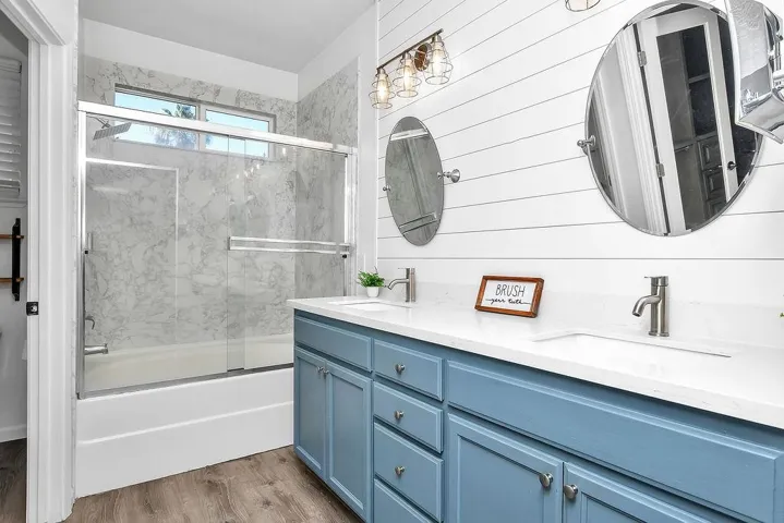 Full bath featuring bath / shower combo with glass door, double vanity, light wood-style flooring, and wooden walls