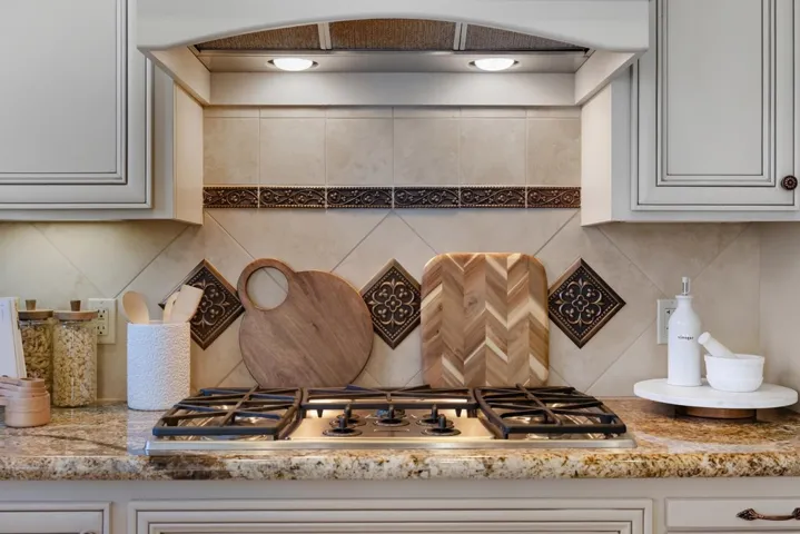 Kitchen view of ventilation hood, tasteful backsplash, stainless steel gas cooktop, and light stone counters