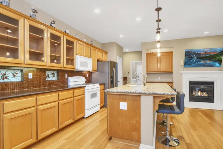 Kitchen featuring white appliances, glass insert cabinets, hanging light fixtures, a kitchen island with sink, and light stone counters