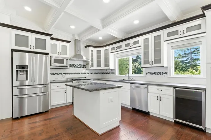 Kitchen featuring stainless steel appliances, white cabinetry, wall chimney exhaust hood, coffered ceiling, and wine cooler