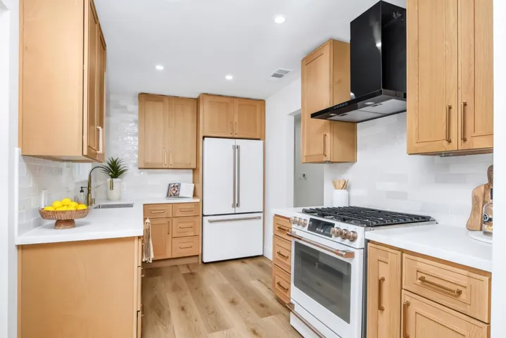 Kitchen featuring white appliances, wall chimney exhaust hood, light wood finished floors, tasteful backsplash, and light brown cabinets