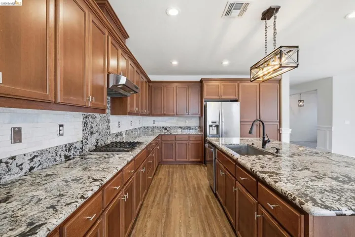 Kitchen featuring dark wood-type flooring, an island with sink, light stone counters, decorative backsplash, and brown cabinetry