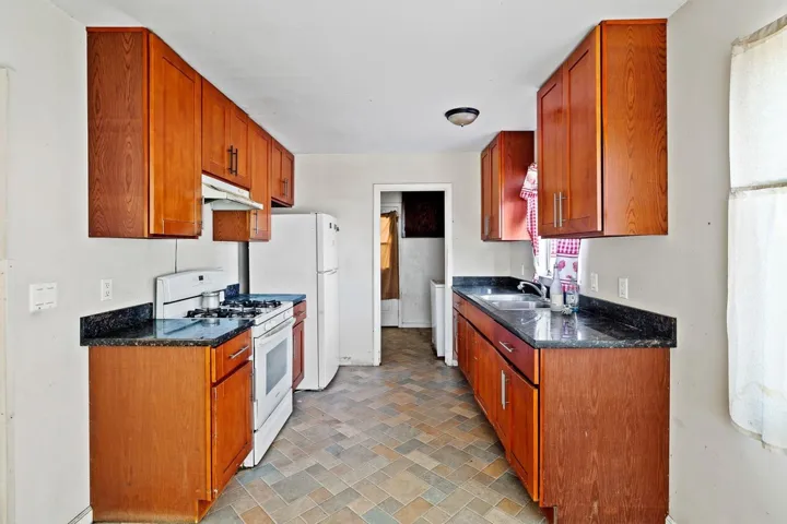 Kitchen with white appliances, brown cabinetry, dark stone countertops, under cabinet range hood, and brick patterned floors