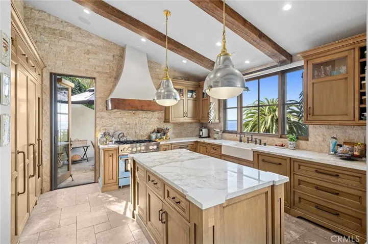 The bespoke kitchen is anchored by a large island and appointed with custom wood cabinetry, polished Carrara marble countertops, and top-tier appliances, all framed by windows overlooking the Pacific Ocean.