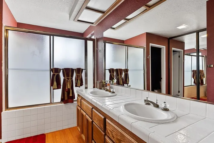 Full bath with a textured ceiling, double vanity, light wood-style flooring, and enclosed tub / shower combo
