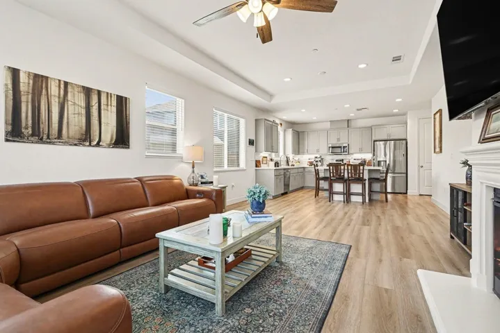 Living room with light wood-style floors, ceiling fan, a fireplace with raised hearth, a raised ceiling, and recessed lighting