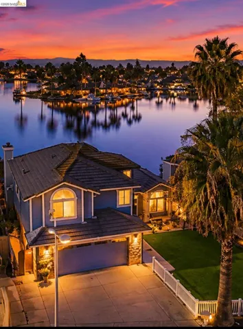 Waterfront property featuring a blue-toned exterior, multi-pane windows, an attached garage, a paved driveway, and a manicured lawn with a white picket fence
