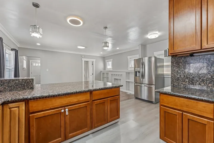 Kitchen featuring wood finish cabinetry, stainless steel fridge with ice dispenser, pendant lighting, ornamental molding, and dark stone countertops