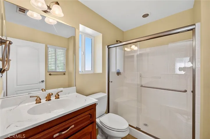 the in-suite bath has a single-sink vanity with solid surface countertop, framed mirror, an a  glass-enclosed shower and natural light from the adjacent window.