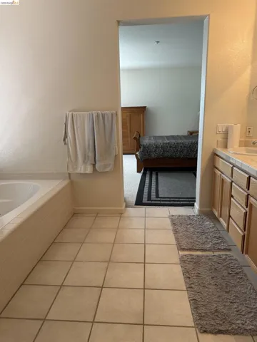 Bathroom featuring a built-in soaking tub with tiled surround, light-toned square floor tiles, and a vanity with light wood-finish cabinetry and a white countertop