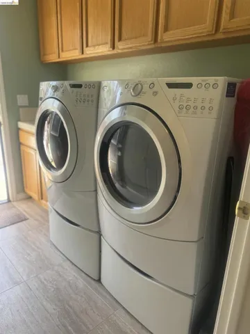 Front-load washer and dryer set on pedestals with a light wood-finish cabinetry overhead and a coordinating countertop
