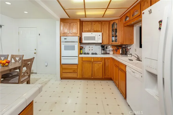 The kitchen is bright and well organized with white tile countertops, a full tile backsplash, and wood cabinetry that extends to the ceiling for abundant storage. A built-in oven, microwave, gas cooktop, and dishwasher provide everyday convenience. The dual basin sink sits beneath a window overlooking the yard.