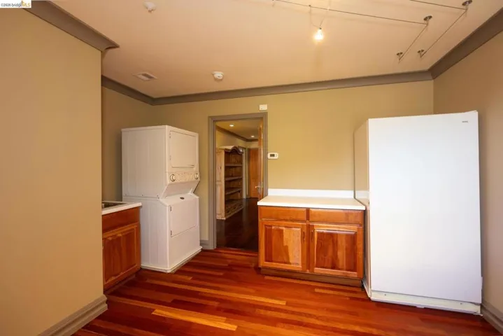 Kitchen with wood finish cabinetry, freestanding refrigerator, stacked washing machine and dryer, light countertops, and dark wood-type flooring