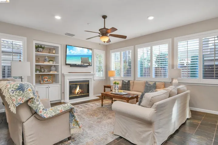 Living area featuring a glass covered fireplace, a ceiling fan, built in shelves, and stone finish flooring
