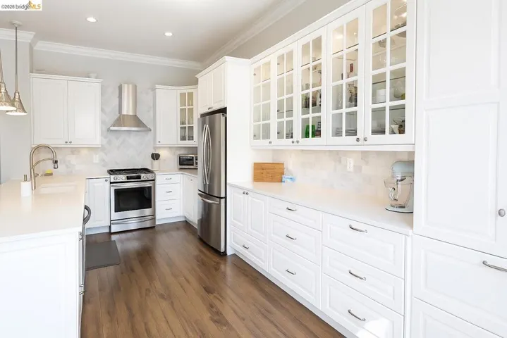 Kitchen featuring decorative light fixtures, appliances with stainless steel finishes, white cabinets, decorative backsplash, and wall chimney exhaust hood