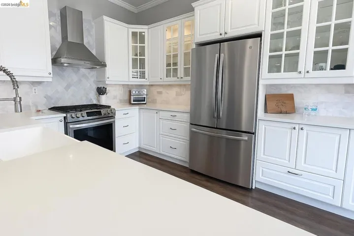 Kitchen featuring decorative backsplash, stainless steel appliances, wall chimney range hood, white cabinetry, and dark wood-style flooring