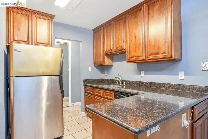 Kitchen featuring freestanding refrigerator, a peninsula, brown cabinetry, dark stone counters, and light tile patterned floors