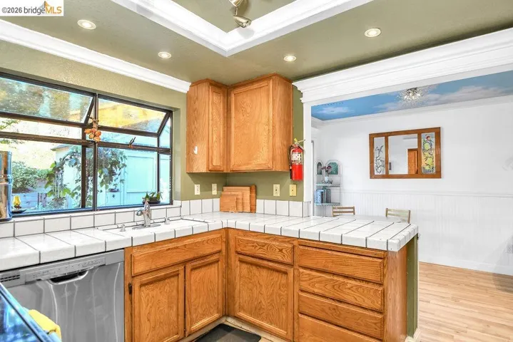 Kitchen with stainless steel dishwasher, a wainscoted wall, tile counters, crown molding, and light wood-style floors