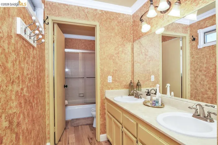 Full bathroom with crown molding, double vanity, bath / shower combo with glass door, wallpapered walls, and light wood-style flooring