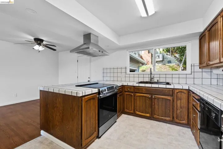 Kitchen featuring tile countertops, a peninsula, backsplash, stainless steel range with electric stovetop, and island range hood