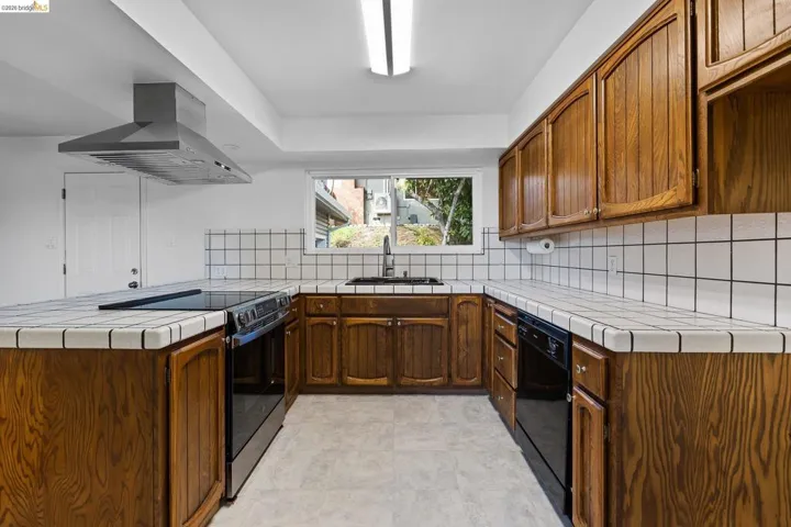 Kitchen featuring tile countertops, a peninsula, black appliances, island range hood, and backsplash