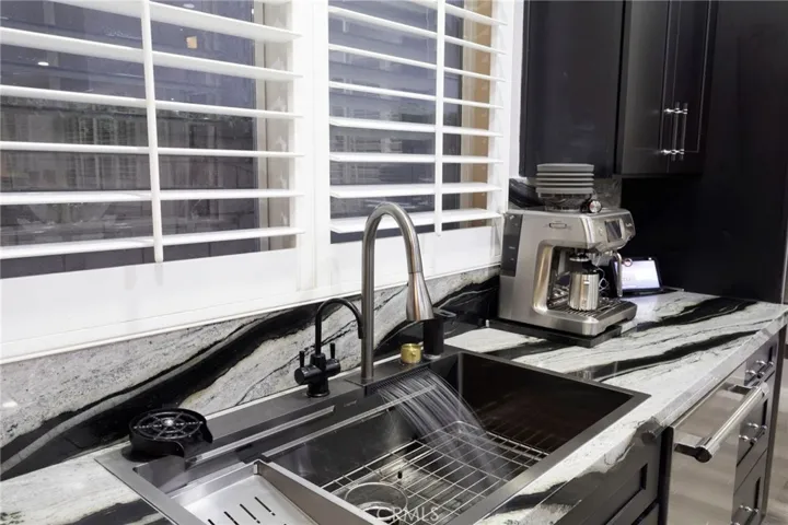 Modern kitchen workspace featuring a large stainless steel workstation sink with integrated accessories, a sleek pull-down faucet, and a secondary filtered water tap. The area is complemented by dramatic quartz countertops with bold waterfall-style veining and matching full-height backsplash, creating a striking designer look. Plantation shutters allow natural light while maintaining privacy, and dark cabinetry with contemporary hardware adds contrast and sophistication.