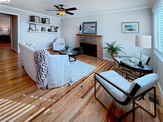 Living room featuring a ceiling fan, light wood finished floors, crown molding, and a brick fireplace