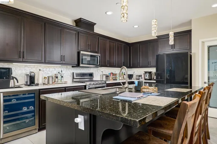 Kitchen with beverage cooler, dark wood finish cabinetry, stainless steel appliances, dark stone countertops, and a center island with sink