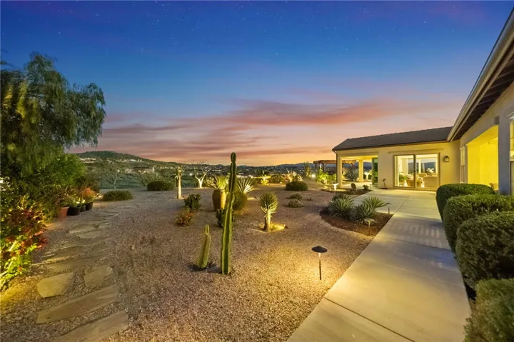 Illuminated desert landscaping and stone pathways create a striking backyard ambiance at dusk with wide-open hillside views.