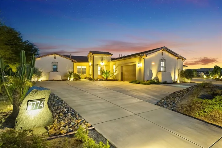 Lighted front elevation highlights the home’s clean lines, generous driveway, and attractive desert landscaping at dusk.