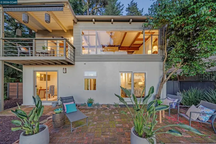 The rear exterior showcases a multi-level layout with a spacious deck, large picture windows, and a lower patio area surrounded by mature landscaping. Sliding glass doors create easy indoor-outdoor flow for relaxing or entertaining.