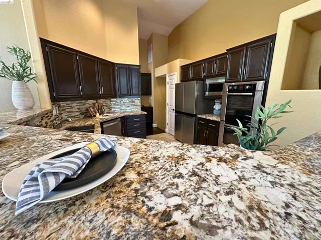 Kitchen featuring light stone counters, stainless steel appliances, backsplash, dark wood finish cabinetry, and light tile patterned floors