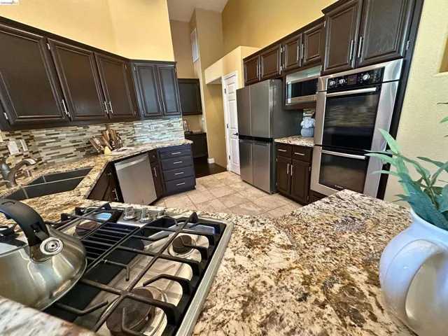 Kitchen featuring stainless steel appliances, light stone counters, dark wood finish cabinetry, a high ceiling, and backsplash