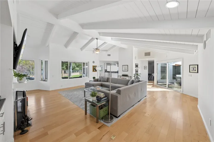 Expansive living area with vaulted beamed ceilings, wide-plank flooring, and oversized windows that bring in abundant natural light. The open layout offers seamless flow to adjacent living spaces and outdoor access.