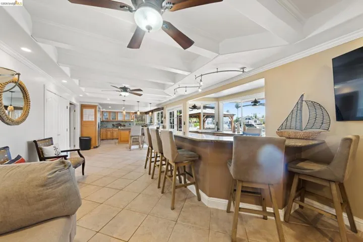 Indoor bar with ornamental molding, beamed ceiling, dark stone counters, light tile patterned floors, and brown cabinetry