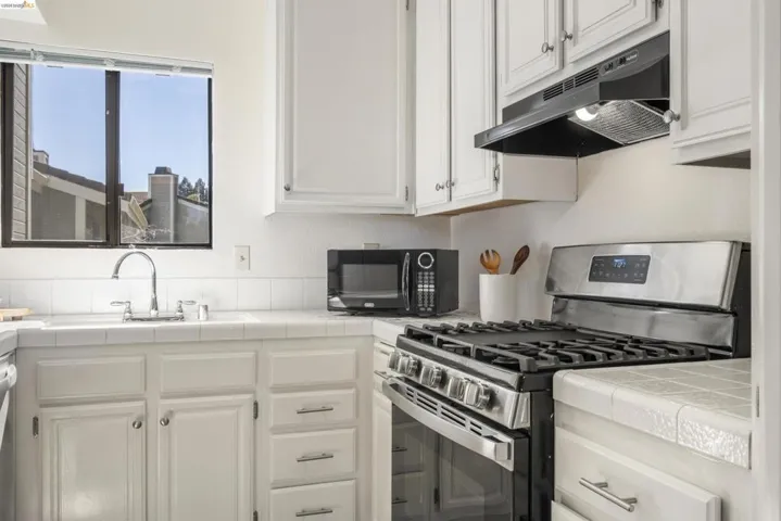 Kitchen featuring stainless steel gas stove, tile counters, white cabinets, and black microwave