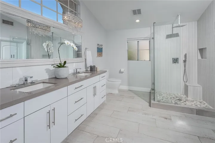 The luxurious primary bathroom offers a spa-inspired setting with a freestanding soaking tub positioned before a large window, a frameless glass-enclosed shower with a waterfall showerhead and handheld fixtures, and a double vanity with storage. High ceilings and beautiful natural light complete the tranquil atmosphere.