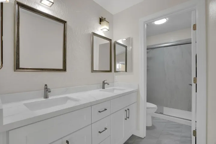 Full bath with double vanity, a marble finish shower, a textured wall, and light marble finish flooring