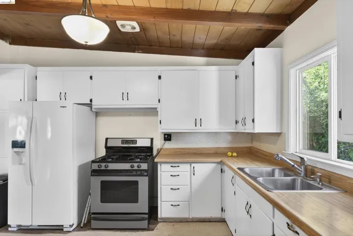 Kitchen with stainless steel gas stove, white cabinets, white fridge with ice dispenser, vaulted ceiling with beams, and light countertops