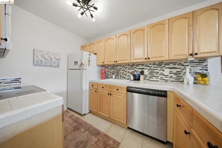 Kitchen featuring tile counters, light wood finish cabinets, dishwasher, and freestanding refrigerator