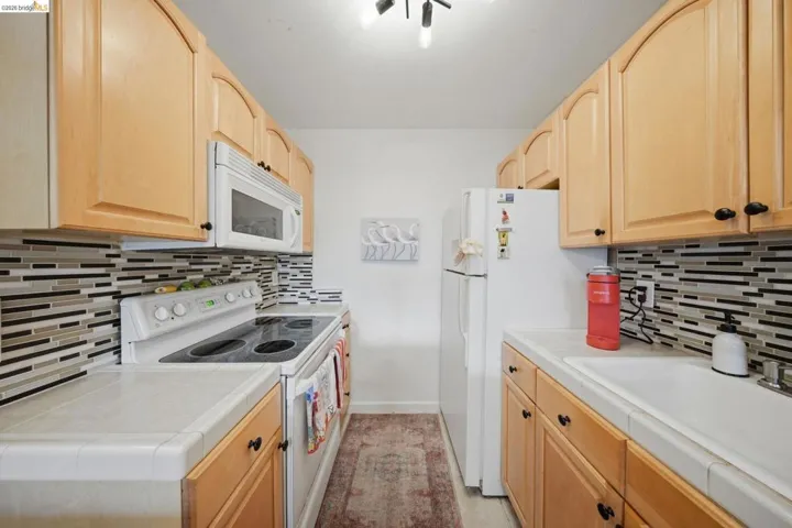 Kitchen with light wood finish cabinetry, white appliances, and tile countertops