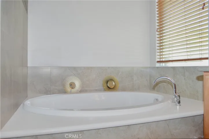 Primary bath with easy-access, walk-in, tiled shower with extra-tall dual shower heads; matching tile surround on soaking tub, dual sinks, Corian-style countertop, wood-look blinds, and tile flooring.