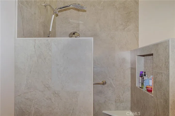 Primary bath with easy-access, walk-in, tiled shower with extra-tall dual shower heads; matching tile surround on soaking tub, dual sinks, Corian-style countertop, wood-look blinds, and tile flooring.
